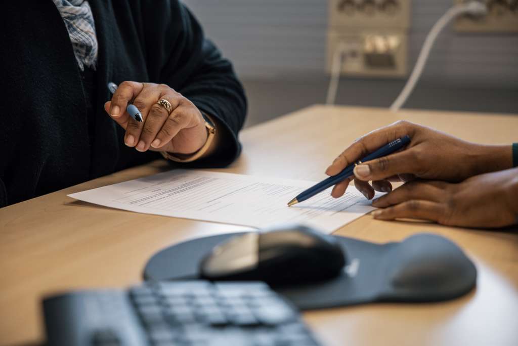 Two people are sitting at a table, both holding a pen in their hands and looking at the same paper. In the foreground of the table, a computer mouse and keyboard are visible.